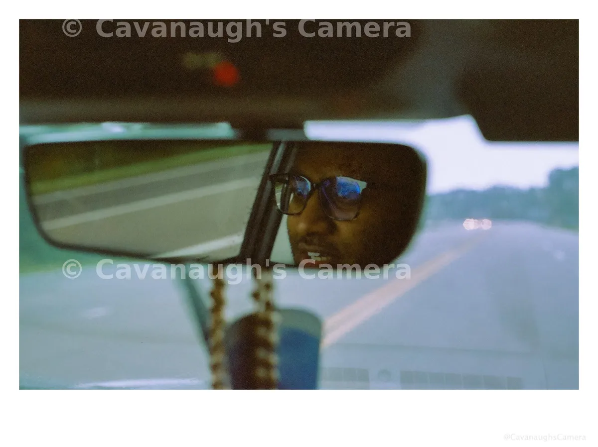 Film self-portrait reflected in a car rearview mirror, wearing glasses with the highway and oncoming headlights behind