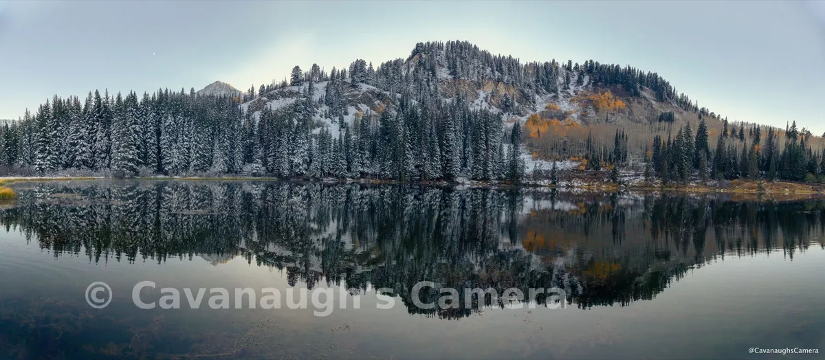 Panoramic view of a snow-dusted mountain ridge with autumn aspens reflected in a still alpine lake