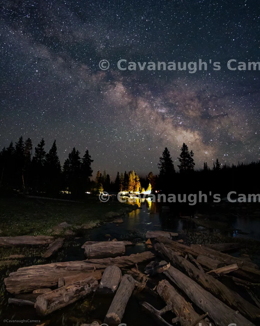 Milky Way arching over a lakeside campground at night, with lit tents glowing among silhouetted pines and driftwood in the foreground