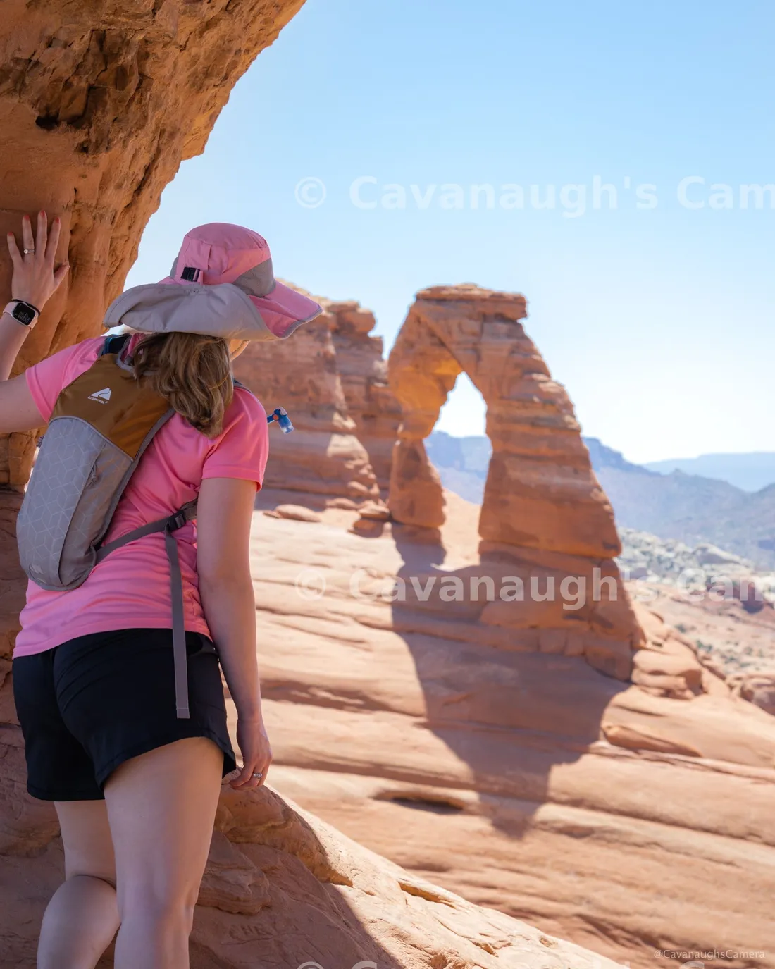 Hiker in a pink shirt and sun hat gazing up at Delicate Arch in Arches National Park