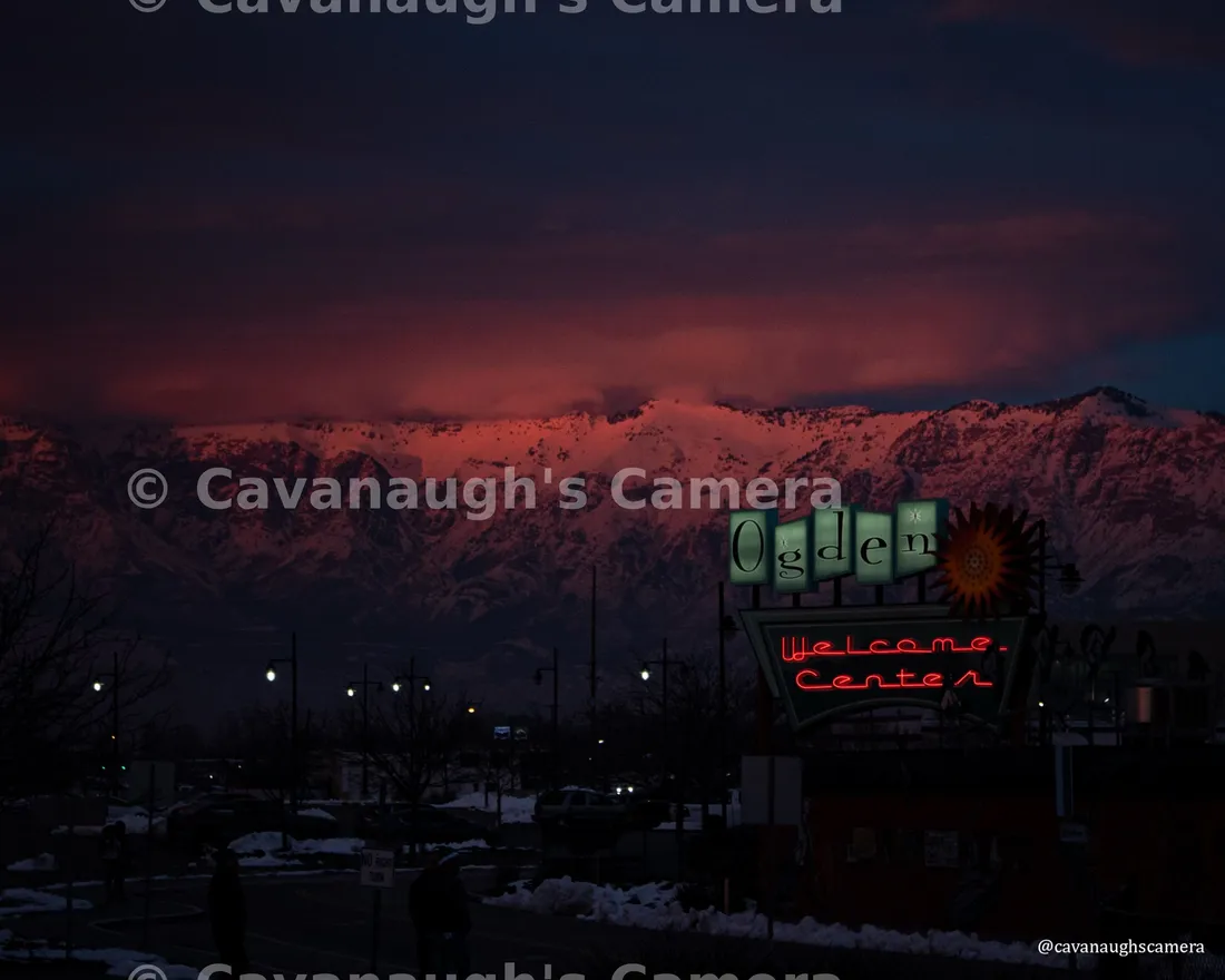 Ogden Welcome Center sign lit up at dusk with snow-covered mountains glowing pink behind it