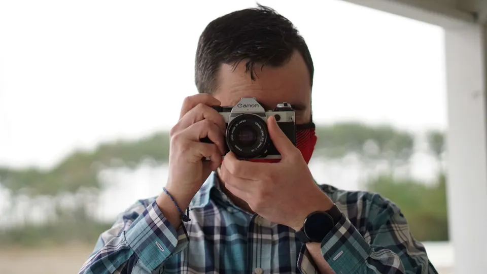 Photographer in a plaid shirt and red bandana holding a vintage Canon film camera up to his eye, blurred greenery behind