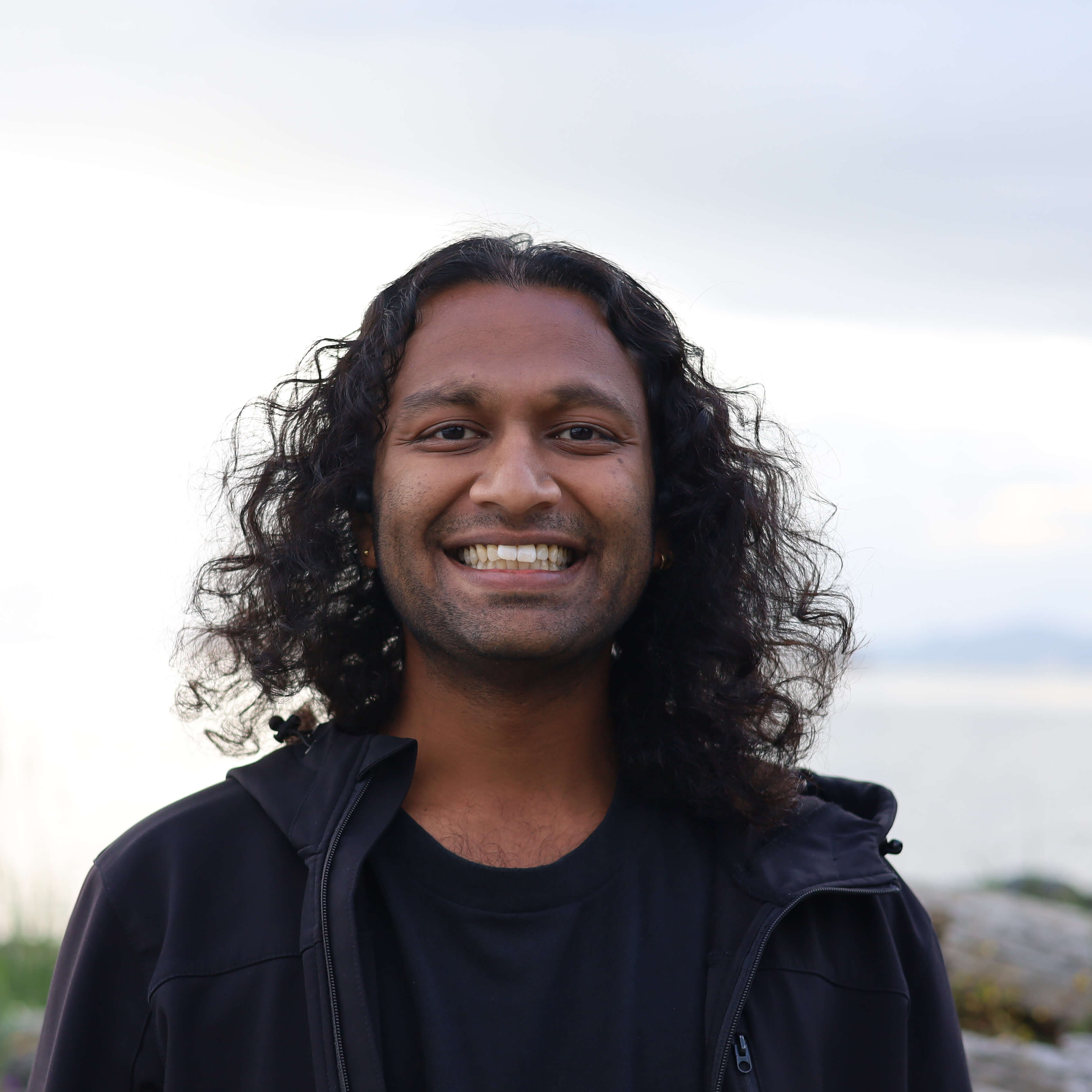 Smiling man with long curly black hair wearing a black jacket outdoors with overcast sky and water in the background